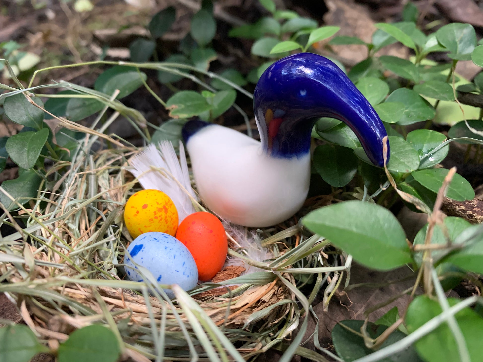 A Whimsical ceramic figure of a Bin Chicken. Blue sitting on a nest of coloured speckled Easter eggs. Bin Chicken in the wild surrounded by green leaves. Rare image.