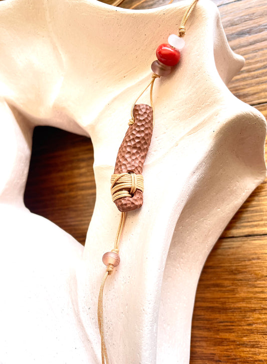 Ceramic pendant with a textured, natural finish, resembling organic patterns found in nature, complemented by a glass bead and hanging from a cord for a rustic yet elegant appearance.