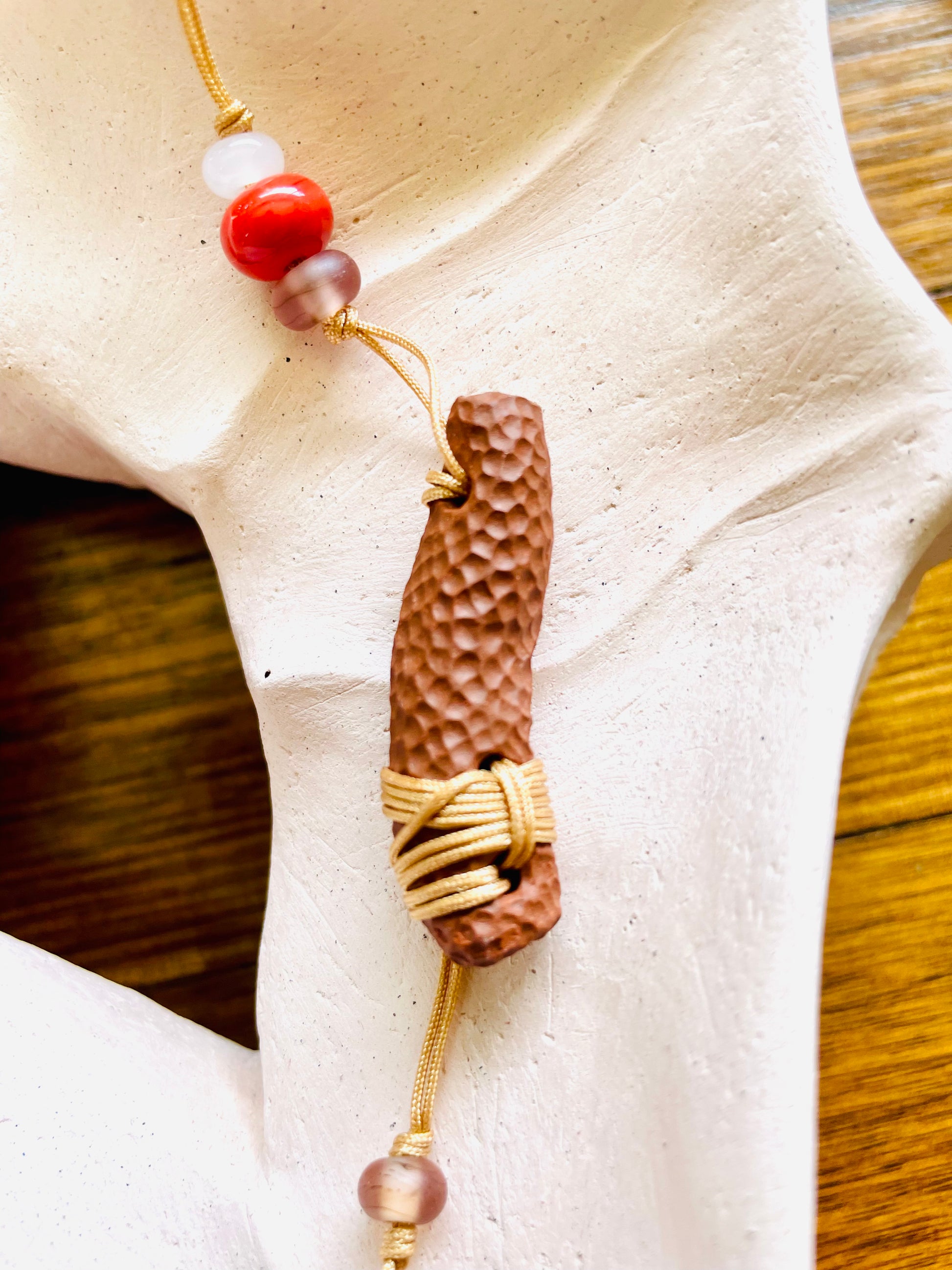 Organic ceramic pendant with a natural texture that mimics the beauty of earthy elements, complemented by glass beads and a gold fabric cord, creating a whimsical and natural accessory.