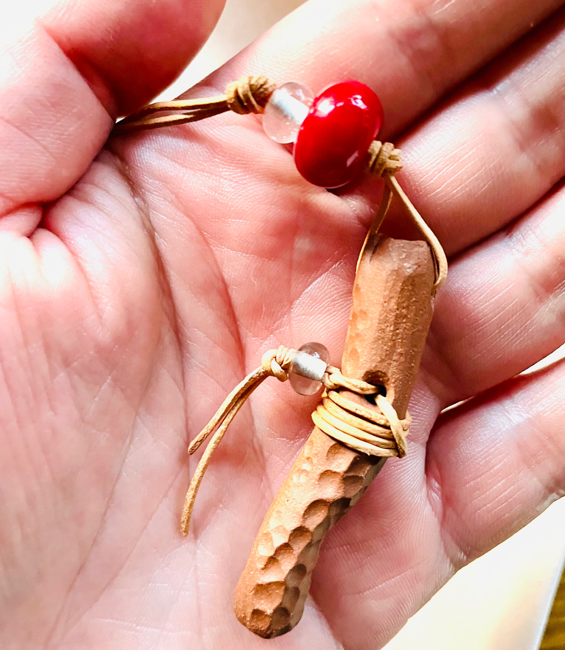Rustic ceramic pendant in a person's hand, adorned with a natural red bead resembling wood and a clear Italian glass bead, showcasing the blend of natural materials and craftsmanship on a light brown leather cord.