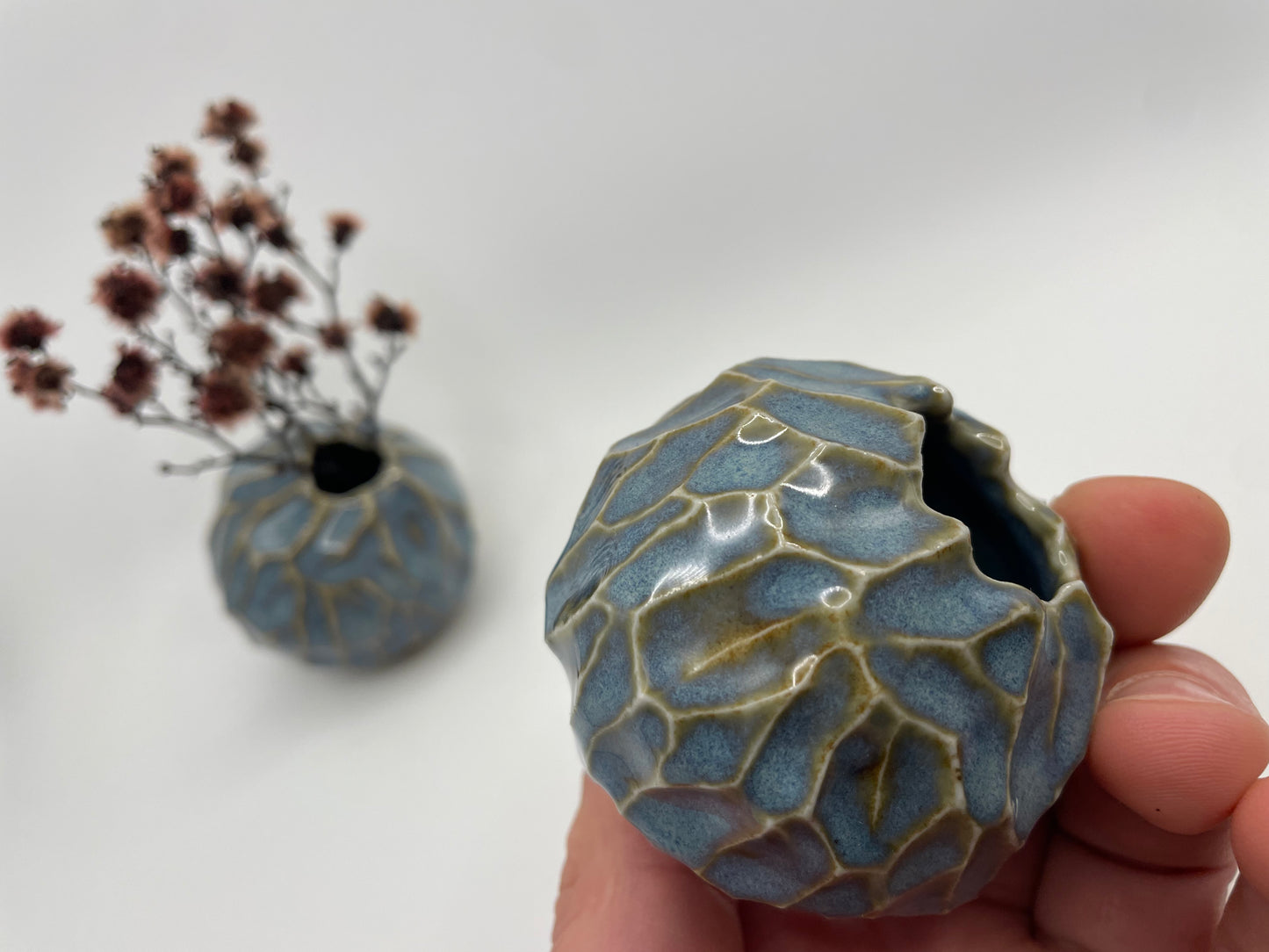 Two blue rutile porcelain vases, one handheld and one in the background, surrounded by a lovely arrangement of dried flowers, highlighting the vases' unique colour and the harmony between the ceramics and the natural elements.