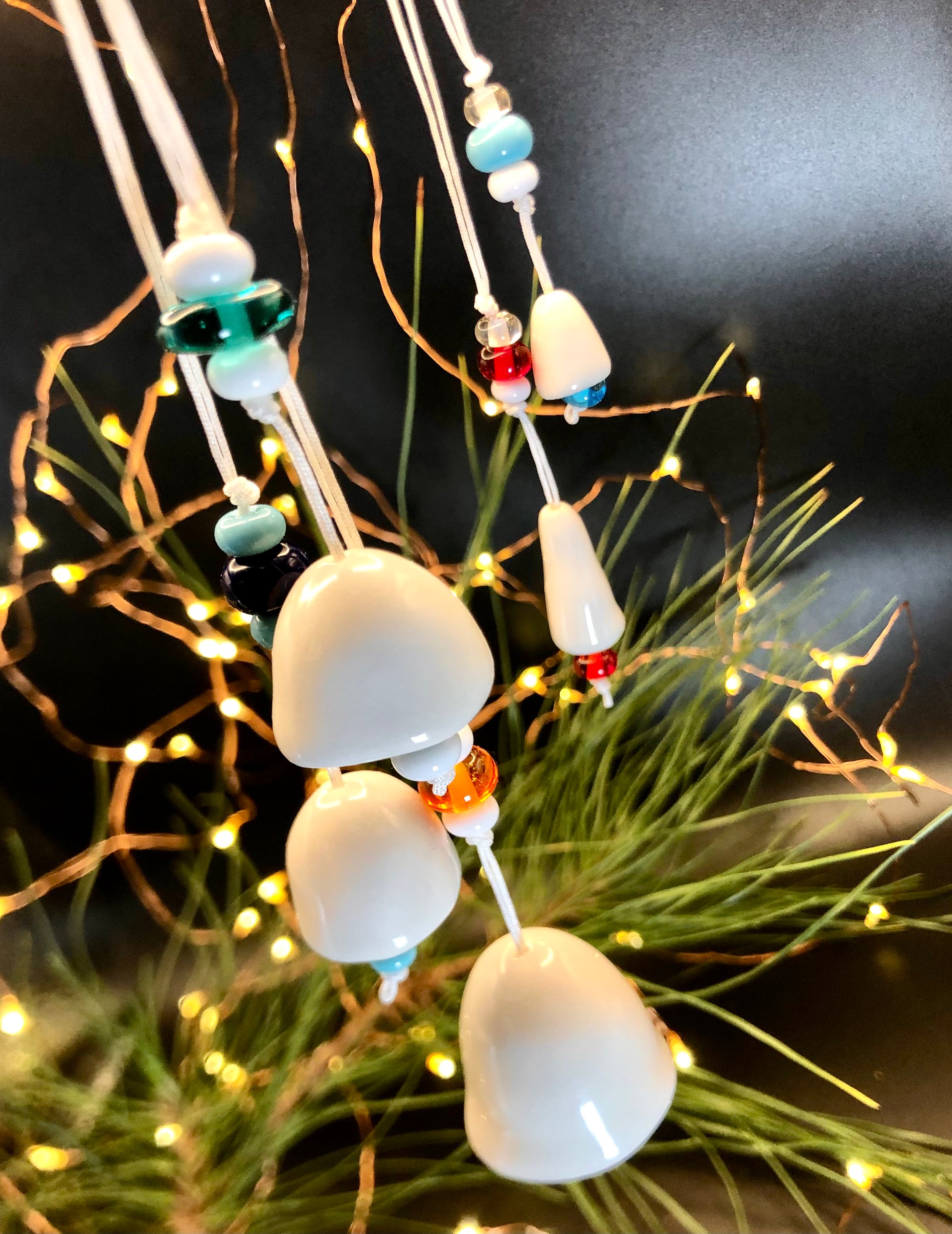 Wonky ceramic bells, adorned with beads, hanging on a Christmas tree amidst twinkling lights, adding a charming and whimsical touch to the festive decorations, blending handmade craftsmanship with the magic of the holiday season.
